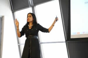 Ritu Bhasin speaking on a conference stage next to a projector screen