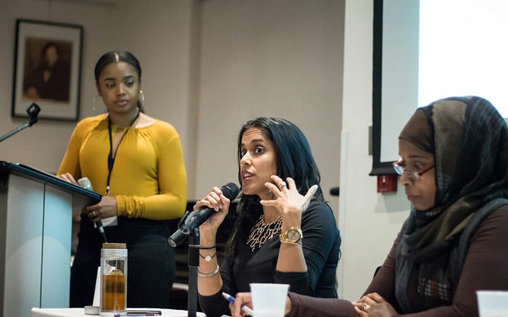 Ritu Bhasin seated at a table speaking into a handheld microphone between a woman standing at a podium and another woman sitting and looking at the table