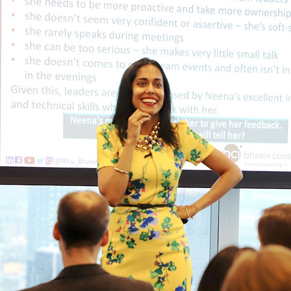 Ritu Bhasin stands in front of a projector screen smiling at a seated crowd while giving a keynote speech