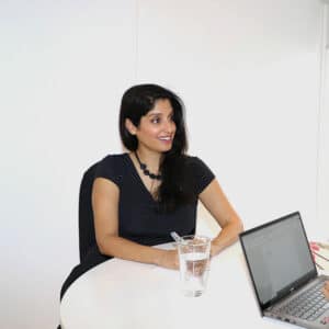Dr. Komal Bhasin sits at a table in a bright white room across from someone with an open laptop smiling at them