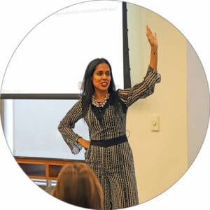 Ritu Bhasin teaching an audience in front of a projector screen with her right hand on her hip and her left hand raised in the air