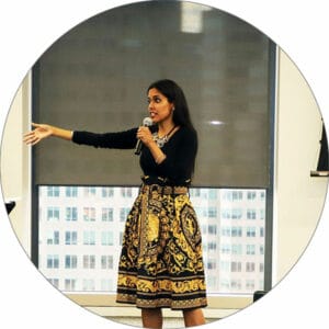 Ritu Bhasin in front of a conference room window speaking into a microphone held in her left hand with her right arm outstreched