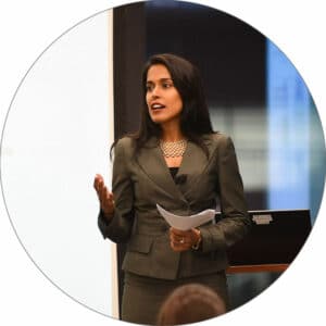 Ritu Bhasin wearing a gray blazer and holding a piece of paper speaking in front of an audience