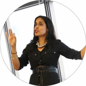 Ritu Bhasin standing in front of a bright window speaking with her right hand raised in front of her and her left arm outstretched