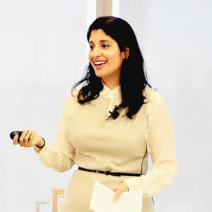 Dr. Komal Bhasin teaching a mental health inclusion workshop in a bright conference room holding a paper in her left hand and a presentation remote in her right hand