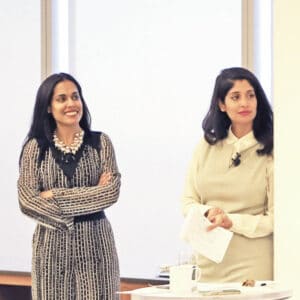 Ritu Bhasin and Dr. Komal Bhasin standing together in a bright conference room looking off to the right. Ritu has her arms crossed and is smiling and Komal looks more serious and holds some notes