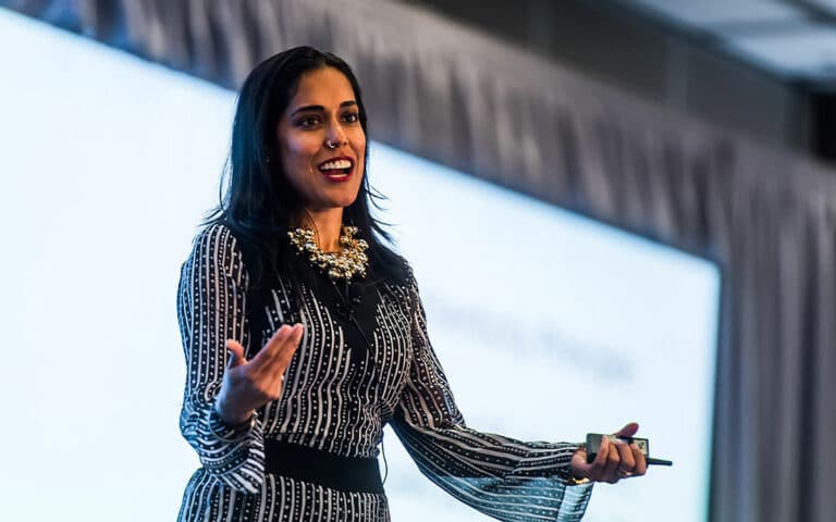 Ritu Bhasin keynote speaking on a stage in front of a projector screen with both hands held in front of her and a remote in her left hand