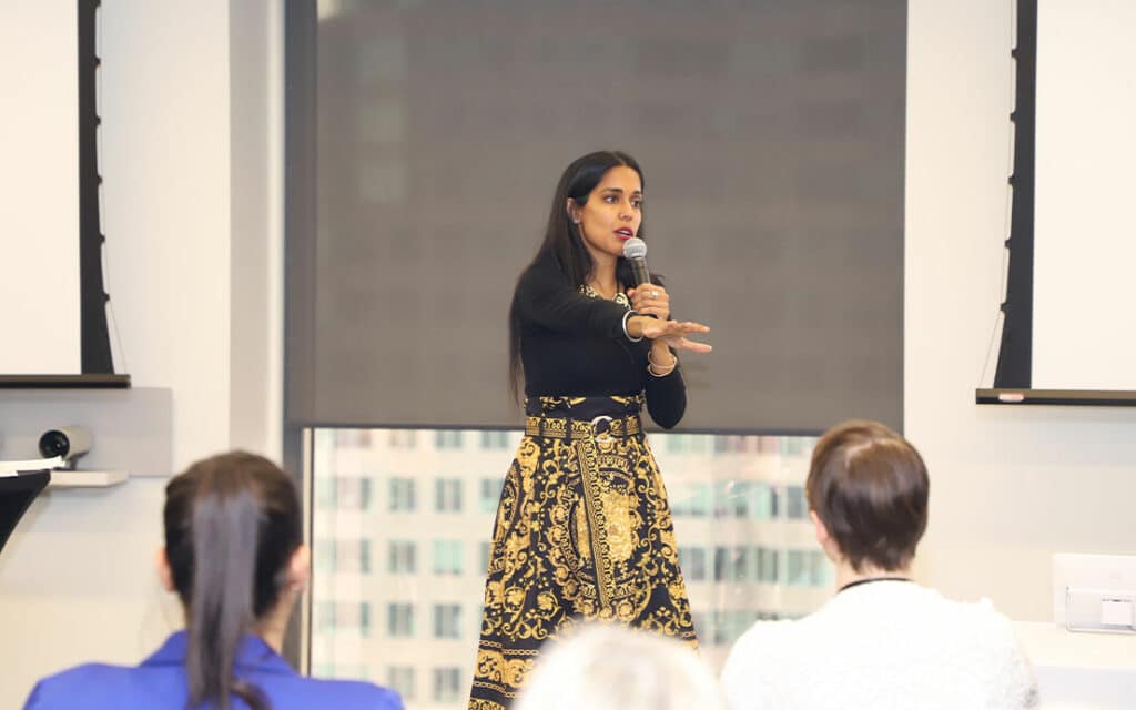 Ritu Bhasin presenting a keynote at the front of a conference room. She holds a microphone and gestures to the crowd in front of her, the backs of whose heads we can see