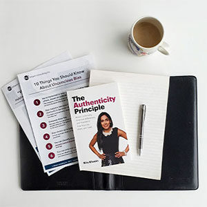 Flat lay of a copy of The Authenticity Principle next to a pen on top of a blank notepad and two bci tip sheets all on top of a black folder on a white table next to a mug of tea