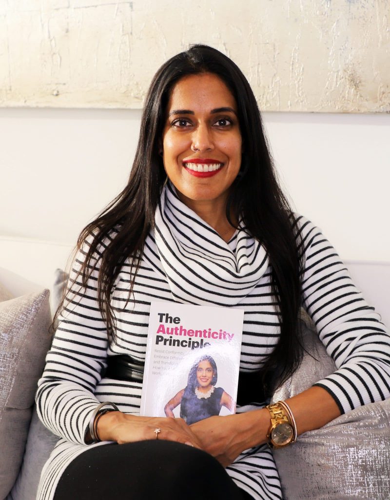 Ritu Bhasin sitting on a couch smiling and holding a copy of her book The Authenticity Principle