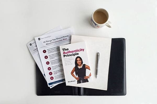 Flat lay of a copy of The Authenticity Principle next to a pen on top of a blank notepad and two bci tip sheets all on top of a black folder on a white table next to a mug of tea