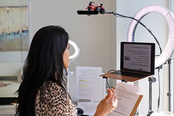 Ritu Bhasin presenting a webinar facing an open laptop with slides onscreen, notes in front of her, and a microphone and ring light